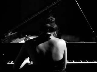 Zdjęcie baneru zakładki "artyści". Czarno białe zdjęcie pianistki grającej przy fortepianie z odkrytymi plecami i związanymi w kok włosami.